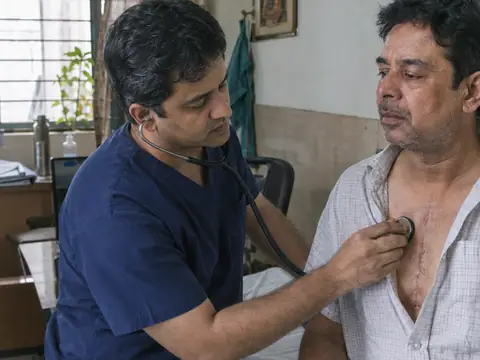 An Indian male cardiothoracic surgeon in navy scrubs listening to the chest of an Indian man patient during a post-operative follow-up visit