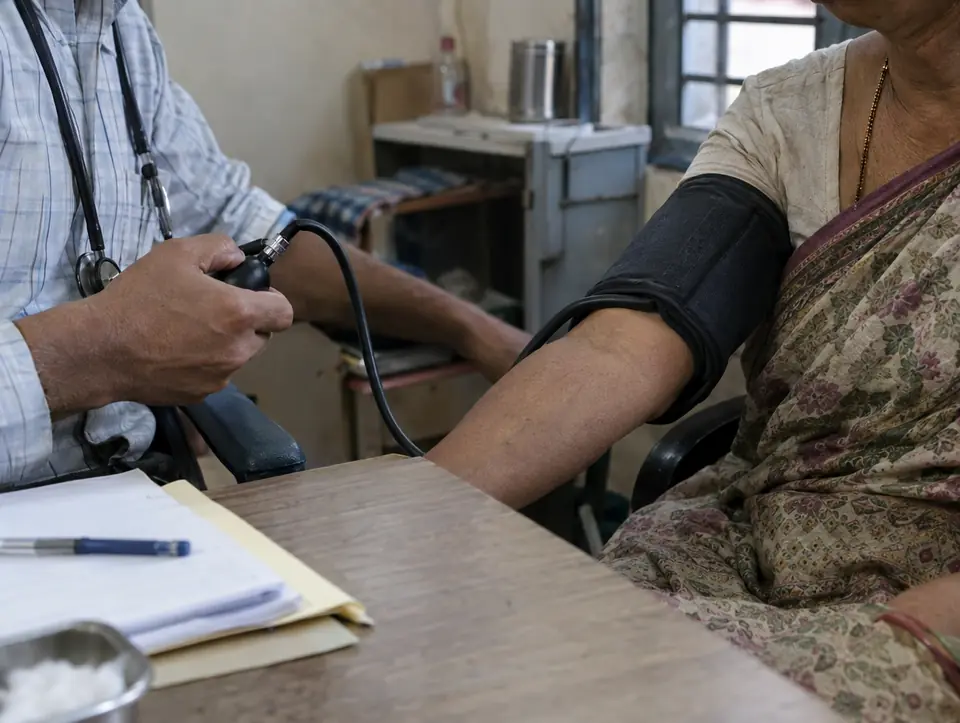 A doctor's hand holding a blood pressure cuff bulb while the cuff is wrapped on an older woman's upper arm over a cotton sleeve, a desk with a pen and paper files