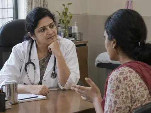 An Indian woman gynaecologist in her forties in a private, gentle conversation with an Indian woman in her thirties across a desk, the patient speaking, the doctor listening