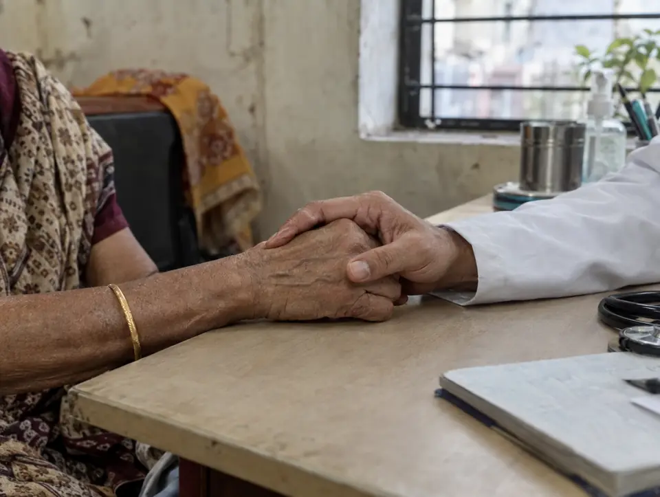 Two hands, one older, one in a white coat sleeve, clasped on a desk