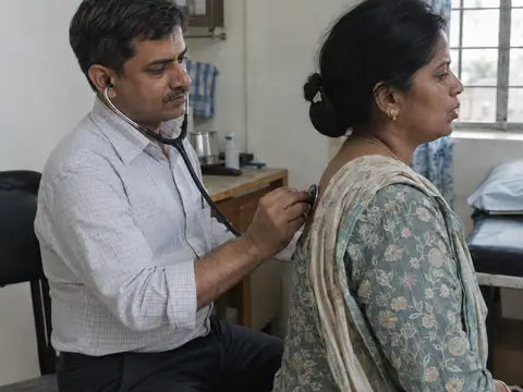 An Indian male chest physician in his forties listening to the back of an Indian woman patient with a stethoscope as she breathes deeply, seated on a stool